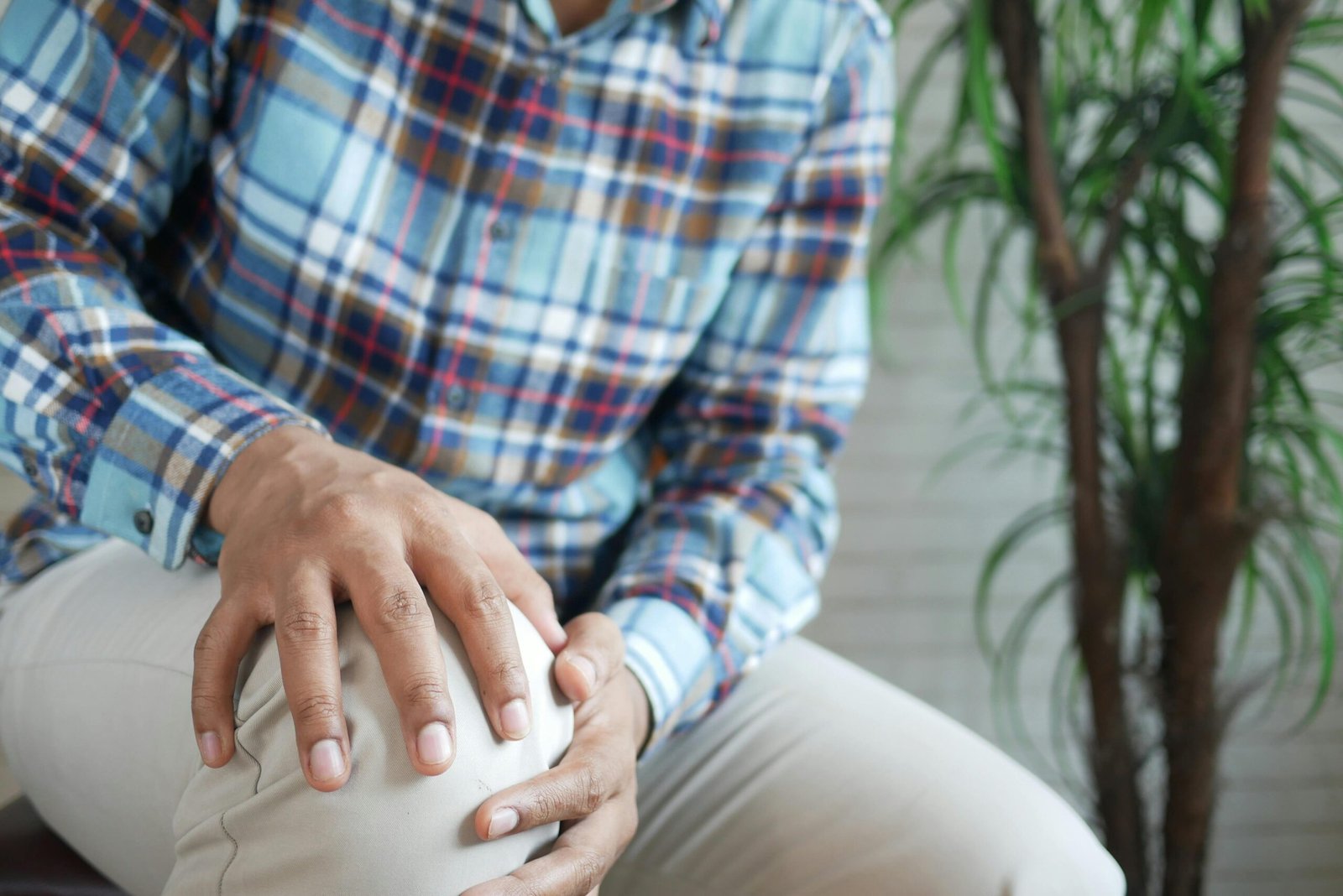 Close-up of a man indoors wearing a plaid shirt, holding his knee, possibly indicating discomfort.